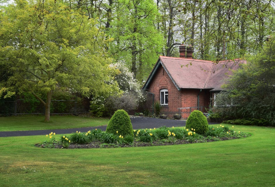 A straight gravel pathway flanked by neatly trimmed grass and evenly spaced wooden posts runs through a lush green garden, with large mature trees on both sides creating a canopy of vibrant foliage. T